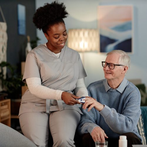 Smiling Black Female Caregiver Using Oximeter to Check Elderly Mans Oxygen Level at Home Supportive Black female social worker visiting upbeat elderly man checking up oxygen saturation in blood with pulse oximeter, while sitting together on couch providing medical assistance at home